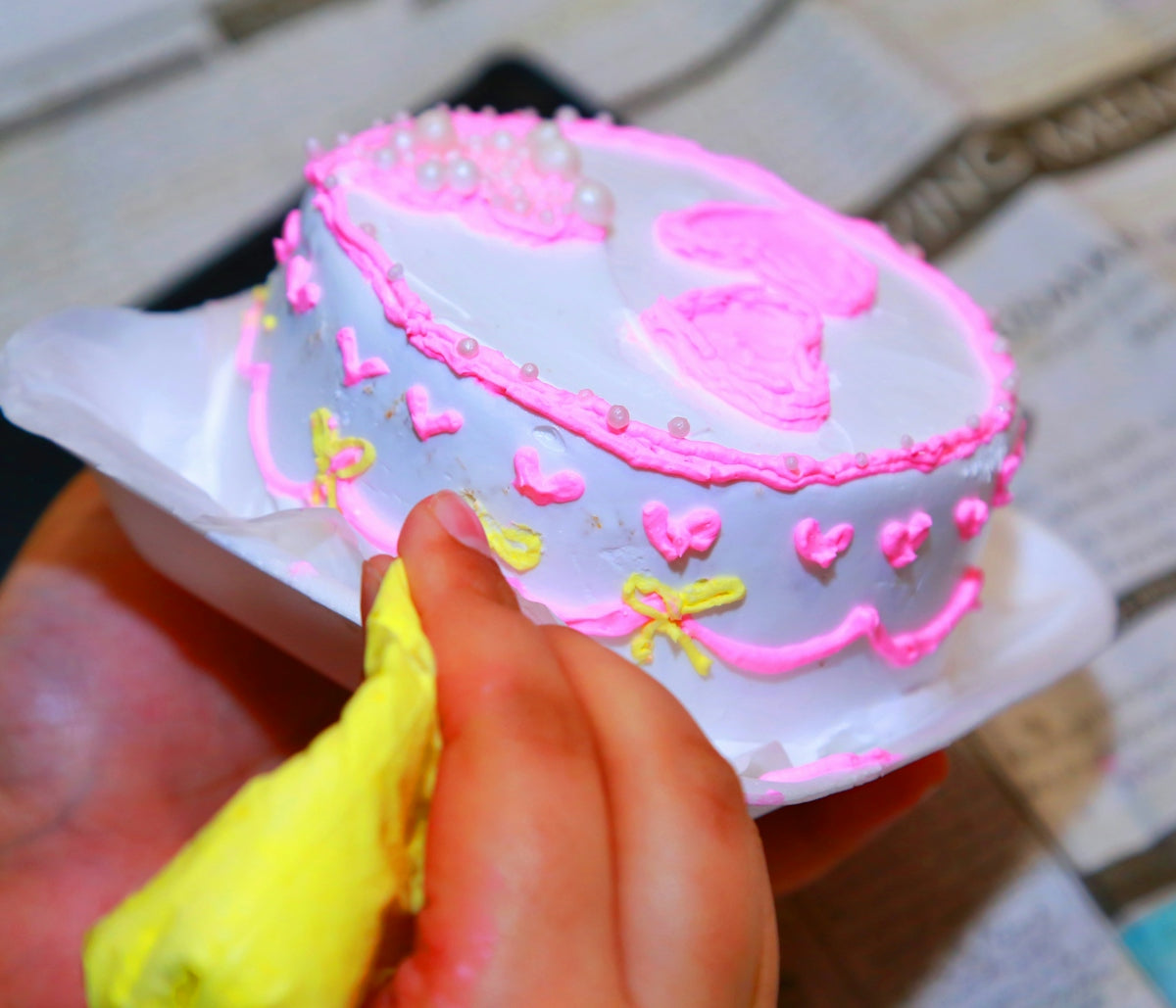 A hand decorating a small cake with pink icing.